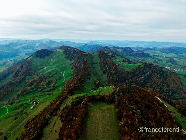 Overlapping ridges of a limestone range receding into blue haze, their crests carrying rust-and-copper autumn forest while broad green pastures spill down the flanks. Pale farm tracks thread between scattered farmsteads, and villages sit in the valleys under a flat, overcast sky. Aerial drone photograph over WASSERFALLEN, Switzerland from 47.370977676820004, 7.719268798828126 on 2025-10-19. Exposure 160 · f1.8 · 9mm.