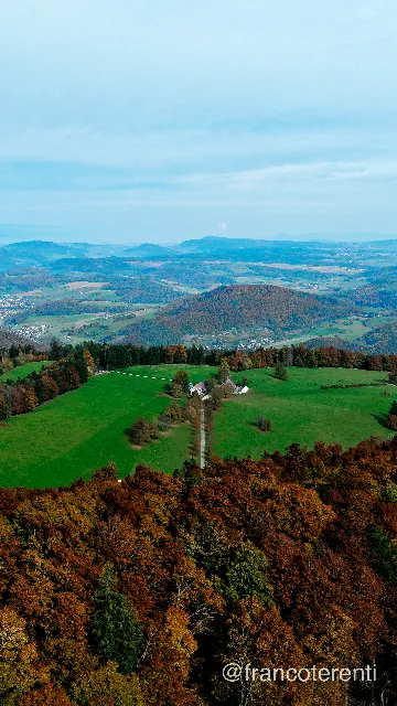 Vertical framing over a shoulder of copper and gold autumn forest, opening onto a plateau of bright green pasture with a farmhouse and a straight track running through it. Beyond, wooded hills and a settled valley fade into layers of blue haze under a pale, thinly clouded sky. Aerial drone photograph over WASSERFALLEN, Switzerland from 47.374872157646564, 7.7160072326660165 on 2025-10-19. Exposure 120 · f1.8 · 9mm.