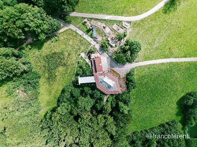 Straight-down view of a hillside station with a dark red hexagonal roof and glazed skylight, an adjoining building with a barn-red roof, and a cable-car track running off to the upper left between trees. A grey road and gravel lanes branch across the frame, a small playground with a blue slide sits beside them, and dense summer woodland meets close-cropped green pasture. Aerial drone photograph over WASSERFALLEN, Switzerland from 47.373724187595116, 7.699785232543945 on 2025-07-22. Exposure 100 · f1.8 · 9mm.
