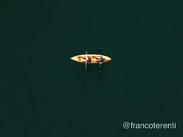 Straight down onto deep green water, empty except for a single yellow tandem kayak near the centre; two paddlers sit with blades raised, casting faint shadows on the calm, faintly rippled surface. Aerial drone photograph over PUNTA NEGRA, Argentina from -31.51504341472417, -68.85766983032228 on 2026-03-03. Exposure 100 · f1.8 · 9mm.