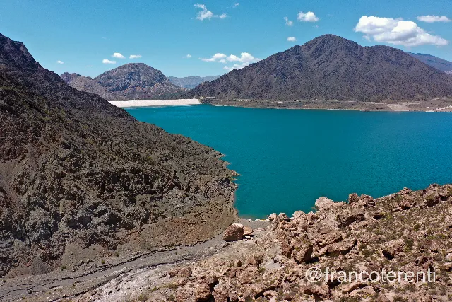 Turquoise reservoir filling a valley between bare, folded brown mountains, with a pale concrete dam wall spanning the far end under a bright blue sky scattered with small white clouds; the rocky, sun-bleached foreground drops steeply to a dry shoreline stripe left by lower water. Aerial drone photograph over PUNTA NEGRA, Argentina from -31.51941697031639, -68.81883144378664 on 2025-01-26. Exposure 100 · f4 · 10mm.