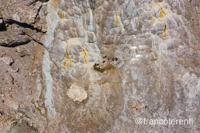 Close overhead look at pale grey and cream mineral terraces streaked with ochre and sulphur-yellow runoff, where a shallow brown pool has formed; a handful of people in swimwear sit and wade at its edge, packs left on the dry rock beside them. Aerial drone photograph over PUNTA NEGRA, Argentina from -31.50941278888475, -68.84114742279054 on 2025-01-26. Exposure 100 · f4.5 · 10mm.