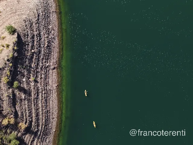 Straight down on a sharp boundary between a dark, dry stony shore ribbed with old waterlines and dotted with scrubby bushes and the deep green water beside it, where a bright band of algae hugs the edge and two small yellow kayaks drift offshore. Aerial drone photograph over PUNTA NEGRA, Argentina from -31.518852650514386, -68.88479232788087 on 2026-03-03. Exposure 100 · f2.8 · 19mm.