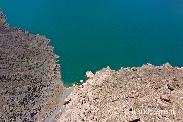 Deep teal water meeting two contrasting shores: dark, sharply eroded grey cliffs and scree on one side, a pale tan boulder field speckled with low dry scrub on the other, all under hard midday sun. Aerial drone photograph over PUNTA NEGRA, Argentina from -31.512304396246748, -68.83127689361574 on 2025-01-26. Exposure 100 · f4 · 10mm.