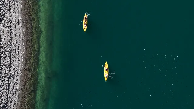 Straight down on two yellow tandem kayaks crossing dark green water, paddles mid-stroke and small wakes trailing behind; along the left edge a grey pebble beach gives way to a wide green band of shallow, algae-covered bottom. Aerial drone video over PUNTA NEGRA, Argentina from -31.519140799914254, -68.8751792907715. Exposure 100 · f2.8.