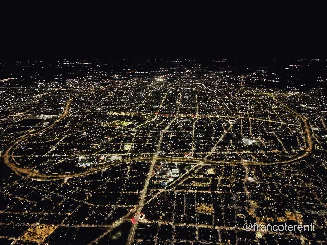 A vast grid of city lights stretching to a black horizon at night, the lit arc of the Circunvalación ring road curving through the lower half and streets glowing amber against dark blocks of low rooftops and unlit tree canopy. Aerial drone photograph over SAN JUAN, Argentina from -31.52982402108432, -68.55520248413087 on 2026-02-24. Exposure 1070 · f1.8 · 9mm.