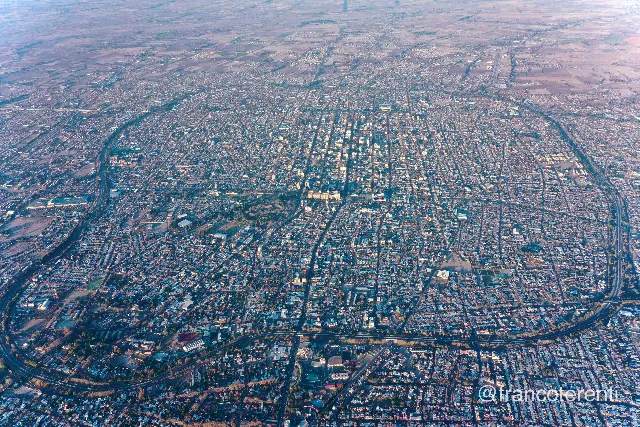 A dense low-rise grid seen from high altitude in cool early light, a compact cluster of taller white buildings marking the centre, the Circunvalación and a rail corridor curving around the edges as tree-lined bands, and pale ploughed fields dissolving into haze at the horizon. Aerial drone photograph over SAN JUAN, Argentina from -31.55791298093802, -68.51503372192384 on 2024-07-25. Exposure 100 · f5 · 10mm.