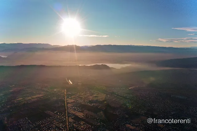 Sunrise over layered mountain ridges, the low sun flaring through dust and haze that fills the valley floor in bands of gold and rose; a reservoir glints in the middle distance above a broad plain of streets, fields and a straight highway running toward the camera. Aerial drone photograph over SAN JUAN, Argentina from -31.5125568213132, -68.52567672729494 on 2024-07-25. Exposure 100 · f6.3 · 10mm.