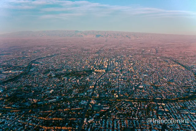 A wide grid of low buildings under a pale blue sky, warm sunset light raking across rooftops and yellowing tree lines, the Circunvalación tracing a green-edged curve around the left and lower edges, with a long arid ridge and dusty plain fading into pink haze on the horizon. Aerial drone photograph over SAN JUAN, Argentina from -31.556011391095595, -68.5455894470215 on 2024-07-25. Exposure 100 · f2.8 · 10mm.