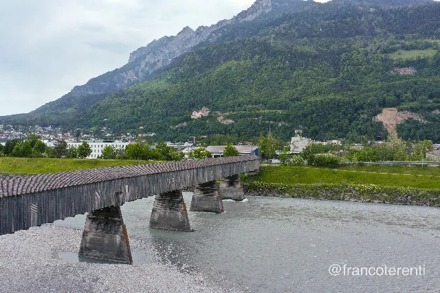 The Old Rhine Bridge, a long covered timber crossing on tapered stone piers, spans a wide pale grey-green river running over gravel banks; low industrial and residential buildings line the far shore beneath a steep forested ridge, with Vaduz Castle small and white on its rocky bluff under flat overcast light. Aerial drone photograph over VADUZ, Liechtenstein from 47.130505495495214, 9.512143135070803 on 2024-05-12. Exposure 100 · f5 · 10mm.
