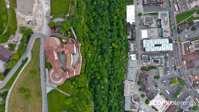 Straight down on Vaduz Castle: red-tiled roofs, square keep, round bastion and curtain walls wrapped around an inner courtyard, ringed by a hairpin road and mown terraces, with a dense wooded scarp separating it from the flat-roofed shops, glass canopies and parking of the Städtle pedestrian centre below. Aerial drone photograph over VADUZ, Liechtenstein from 47.14050440064906, 9.521552324295046 on 2024-05-12. Exposure 100 · f4.5 · 10mm.