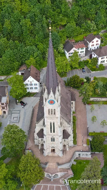 The Cathedral of St. Florin seen at a steep angle: grey stone Gothic Revival nave and tall dark spire with a gilded finial and blue clock face, rising from a paved terrace of steps and low walls among white shuttered houses, lawns and heavy deciduous woodland. Aerial drone photograph over VADUZ, Liechtenstein from 47.13630800882749, 9.52246427536011 on 2024-05-12. Exposure 100 · f4.5 · 10mm.
