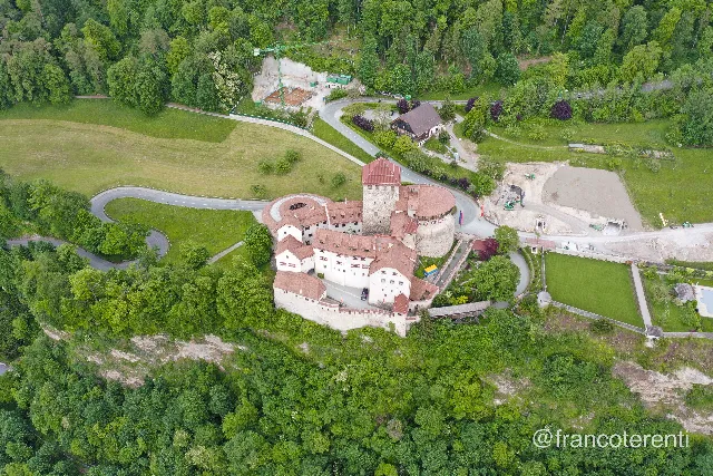 Vaduz Castle on its wooded rocky bluff — white walls, red tile roofs, square keep, round bastion and crenellated curtain walls — encircled by a hairpin road, sloping green meadows and mature trees, with a construction site, crane and bare earth on the plateau behind. Aerial drone photograph over VADUZ, Liechtenstein from 47.13934404021297, 9.523719549179077 on 2024-05-12. Exposure 100 · f4 · 10mm.