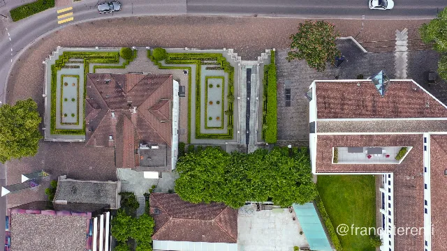 Straight down on a red-tiled hipped-roof villa flanked by symmetrical clipped-hedge parterres and two shallow rectangular basins, with broad paved plazas, conical topiary and a stepped ramp; the black cube of the Kunstmuseum Liechtenstein, its glass band shot with pink, sits at the lower left among leafy trees. Aerial drone photograph over VADUZ, Liechtenstein from 47.13615474303139, 9.519621133804321 on 2024-05-12. Exposure 100 · f4.5 · 10mm.