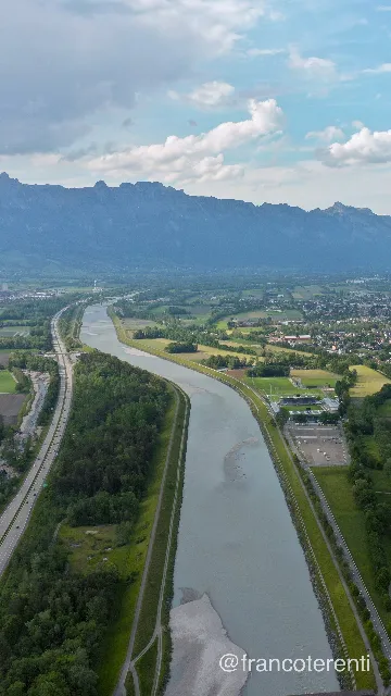 The channelled Rhine, silt-pale with exposed gravel bars, runs straight between raised embankments down a wide green valley — motorway and woodland belts on one side, the Rheinpark Stadion, its training pitches and car park, farm plots and a spread-out town on the other — backed by a long hazy blue mountain wall under scattered cumulus. Aerial drone photograph over VADUZ, Liechtenstein from 47.137570610242776, 9.510040283203127 on 2024-05-12. Exposure 100 · f5.6 · 10mm.