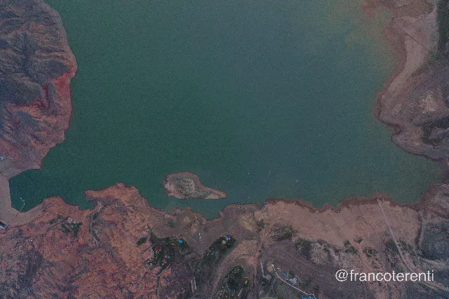 Top-down over deep teal water framed by bare, rust-red rock ridges and pale eroded flats; a small crescent island sits mid-frame, while the lower shoreline shows sandy beaches, scattered cabins with bright roofs, dirt tracks and a jetty under flat, cool light. Aerial drone photograph over ULLUM, Argentina from -31.468972225704622, -68.66073131561281 on 2024-10-19. Exposure 400 · f2.8 · 10mm.