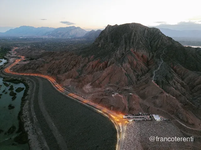 Dusk over a dark ridge of layered grey and red rock rising above dry badlands, with a curving highway traced by orange headlight streaks along a long embankment, parked cars and a lit stall at the roadside, shallow water and sandbars on the left and hazy blue ranges on the horizon. Aerial drone photograph over ULLUM, Argentina from -31.47933043844773, -68.65772724151613 on 2026-03-02. Exposure 330 · f1.8 · 9mm.