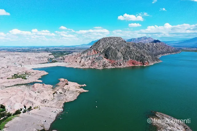 Turquoise reservoir under a bright blue sky with scattered cumulus, hemmed by a stark grey mountain streaked with red rock strata on one shore and pale, eroded sand-coloured badlands and low bluffs on the other; small boats float near a shoreline beach dotted with parked vehicles. Aerial drone photograph over ULLUM, Argentina from -31.466483155717043, -68.6695718765259 on 2025-03-02. Exposure 100 · f4.5 · 10mm.