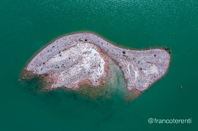 Straight down onto a small elongated island of pale grey and pinkish rubble speckled with low green shrubs, ringed by submerged boulders and a shallow jade halo that fades into flat deep-green open water; a single red kayak drifts at the right edge. Aerial drone photograph over ULLUM, Argentina from -31.465970691916294, -68.6565685272217 on 2025-03-02. Exposure 100 · f4 · 10mm.