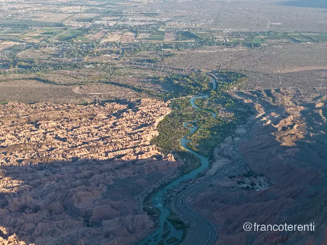 Low sun rakes across deeply furrowed, pinkish-tan badlands that drop to a narrow valley where a turquoise river meanders through bright green riverside scrub, a paved road curving alongside it and irrigated fields and a spread-out town filling the hazy plain beyond. Aerial drone photograph over ULLUM, Argentina from -31.45868608173212, -68.66253376007081 on 2026-02-28. Exposure 100 · f2.8 · 19mm.