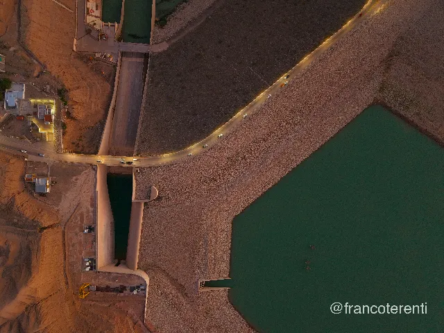 Top-down at dusk on a concrete dam and spillway works, with a warm-lit road curving across the rockfill crest, headlights and taillights of passing cars, floodlit service buildings on the bare orange hillside and calm dark-green reservoir water filling the right of the frame. Aerial drone photograph over ULLUM, Argentina from -31.474499188582687, -68.64961624145509 on 2026-03-02. Exposure 270 · f1.8 · 9mm.