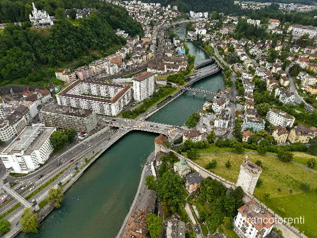 The green-tinted Reuss curves between steep wooded slopes and packed apartment blocks, passing under the Geissmattbrücke and further road and rail crossings; the squat round Nölliturm anchors the water's edge, while the Museggmauer climbs the mown lawn on the right to the battlemented Männliturm and the turreted Château Gütsch crowns the trees upper left. Aerial drone photograph over LUCERNE, Switzerland from 47.054510846333585, 8.294591903686525 on 2026-06-12. Exposure 110 · f1.8 · 9mm.