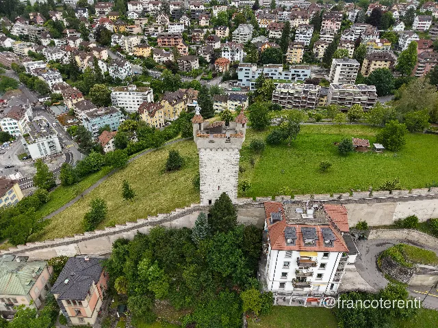 The whitewashed Männliturm rises from a grassy ridge, its red-tiled cap topped by the small armoured figure that gives it its name, with the crenellated Museggmauer running downhill past mature trees and pastel townhouses and balconied apartment blocks stacked on the slope beyond. Aerial drone photograph over LUCERNE, Switzerland from 47.053224344717066, 8.301630020141603 on 2026-06-12. Exposure 100 · f1.8 · 9mm.