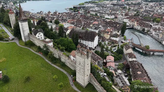 The Museggmauer runs along a green hillside with its square battlemented towers — the round Nölliturm and spired Männliturm at the left, a taller keep in the foreground — above the tight tiled roofs of the Altstadt, with the covered Spreuerbrücke, a slender steel footbridge over the Reuss weir and the Jesuitenkirche domes on the right under flat overcast light. Aerial drone video over LUCERNE, Switzerland from 47.054598561222726, 8.306393623352053. Exposure 100 · f2.8.
