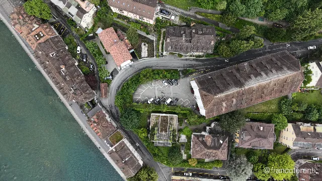Straight-down framing of tiled roofs and a stone quay wall along the milky turquoise Reuss, with a road curling around a small car park of marked bays and parked cars, a long dark-roofed hall alongside, and clipped hedges, garden trees and a rooftop terrace filling the gaps. Aerial drone video over LUCERNE, Switzerland from 47.05591426724654, 8.300814628601076. Exposure 100 · f2.8.