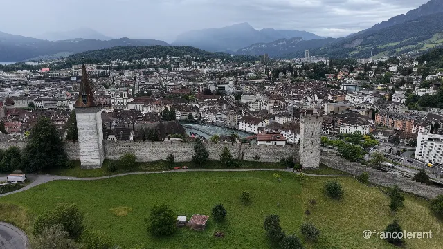 The spired Männliturm and a second battlemented tower anchor the crenellated Museggmauer across a mown hilltop with scattered trees and a small shed; below spread the tiled roofs and church steeples of the Altstadt along the green Reuss, backed by forested slopes and layered blue peaks under low cloud. Aerial drone video over LUCERNE, Switzerland from 47.050154158639906, 8.299913406372072. Exposure 100 · f2.8.