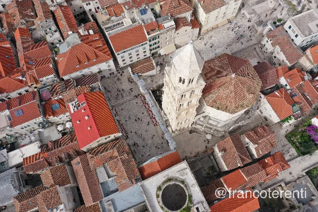 Straight-down look at the Cathedral of Saint Domnius in Split, its pale stone bell tower and octagonal tiled dome rising from the dense terracotta rooftops of Diocletian's Palace, with crowds and rows of red chairs set out on the Peristyle below and the circular walled Vestibule courtyard in the foreground. Aerial drone photograph over SPLIT, Croatia from 43.51074740953776, 16.436190605163578 on 2024-06-21. Exposure 100 · f5.6 · 10mm.