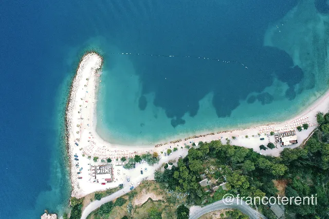 Overhead frame of Kašjuni beach on the Marjan peninsula, a white pebble spit curving into deep blue and turquoise water, edged with rock armour and lined with parasols, sunbeds and parked cars, backed by the pine-covered slope of Marjan Forest Park. Aerial drone photograph over SPLIT, Croatia from 43.500658802113065, 16.439967155456547 on 2024-06-21. Exposure 200 · f2.8 · 10mm.