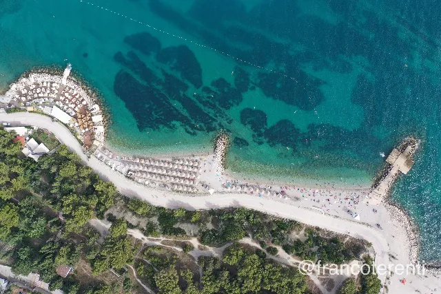 Top-down view of the Kašjuni shoreline below Marjan hill, its curved pebble beach packed with beige parasols and bathers, backed by the seafront road, dry scrub and dense pines, with dark seagrass patches mottling the clear turquoise shallows offshore. Aerial drone photograph over SPLIT, Croatia from 43.50314932396644, 16.443185806274418 on 2024-06-20. Exposure 100 · f3.5 · 10mm.