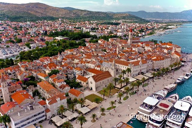 Low oblique over the stone old town of Trogir, the slender campanile of St Lawrence's Cathedral standing above orange tile roofs, with large white yachts moored along the palm-lined Riva and low green hills fading into haze under bright daylight. Aerial drone photograph over SPLIT, Croatia from 43.50271340666097, 16.42636299133301 on 2024-06-23. Exposure 100 · f3.2 · 10mm.
