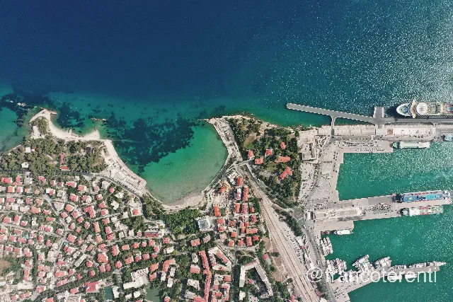 Straight-down composition splitting Split's ferry port — long concrete piers, ferries and a moored cruise ship — from the leafy hillside of red-roofed villas above Bačvice, where a milky turquoise bay and its shallow sandy beach fill the left of the frame. Aerial drone photograph over SPLIT, Croatia from 43.49910127068052, 16.43541812896729 on 2024-06-22. Exposure 100 · f5.6 · 10mm.