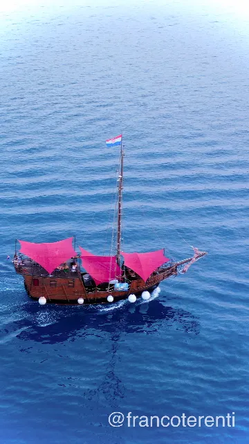 Wooden replica galleon of the kind that runs excursions along the Dalmatian coast, three magenta sails set and the Croatian flag at the masthead, white fenders along her hull as she cuts a thin wake across rippled deep blue open water. Aerial drone photograph over SPLIT, Croatia from 43.509405720754536, 16.42662048339844 on 2024-06-21. Exposure 100 · f3.2 · 10mm.