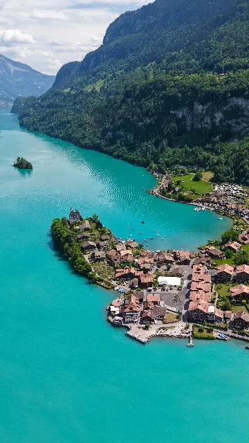 Tall vertical framing down a turquoise lake, with a village of brown and terracotta roofs and a turreted mansion occupying a wooded peninsula in the foreground, boats moored in the sheltered bay, and steep forested cliffs falling to the water beside a small treed islet. Aerial drone photograph over ISELTWALD, Switzerland from 46.7129150888077, 7.959959506988526 on 2026-05-31. Exposure 120 · f1.8 · 9mm.