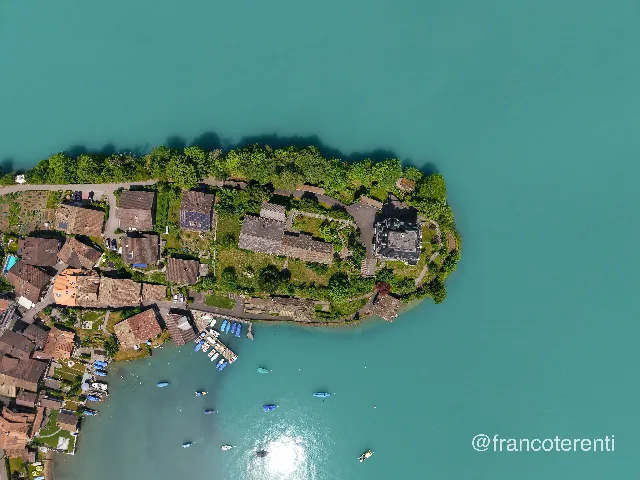 Straight-down look at a wooded peninsula tipping into flat turquoise water, its tree-covered spine ringed by tiled roofs, terraced gardens and a dark slate-roofed mansion; a small marina of jetties and moored boats sits at the lower left, with sunlight glinting off the shallows. Aerial drone photograph over ISELTWALD, Switzerland from 46.71288452230209, 7.965259552001954 on 2026-05-31. Exposure 100 · f1.8 · 9mm.