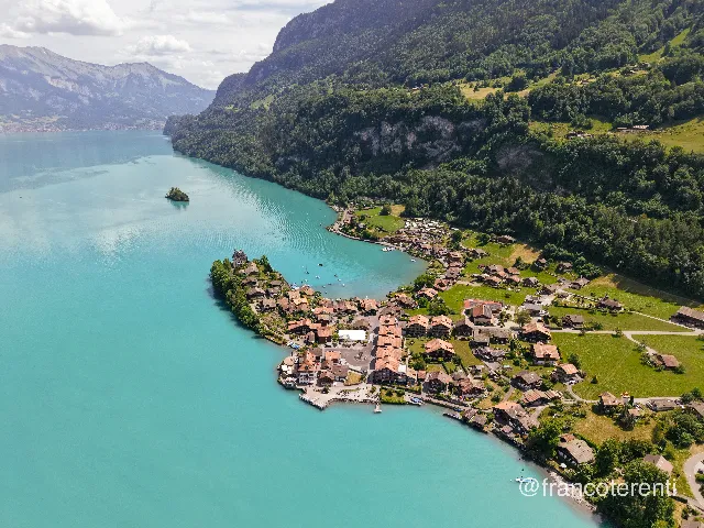 A lakeside village of orange-tiled chalets spreads across a green headland where a turquoise lake meets steep forested slopes and grey limestone cliffs; a tiny wooded islet floats offshore and hazy blue peaks close the far end of the valley under a bright, partly clouded summer sky. Aerial drone photograph over ISELTWALD, Switzerland from 46.710928355686974, 7.959830760955811 on 2026-05-31. Exposure 120 · f1.8 · 9mm.