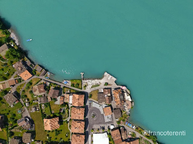 Overhead frame of a shoreline settlement where a small square, a fountain roundabout and clustered terracotta roofs meet a stone quay, a boat landing and rows of moored dinghies, with pale turquoise water filling the upper half and a single motorboat cutting a wake near the beach. Aerial drone photograph over ISELTWALD, Switzerland from 46.711810630653765, 7.9626202583313 on 2026-05-31. Exposure 100 · f1.8 · 9mm.