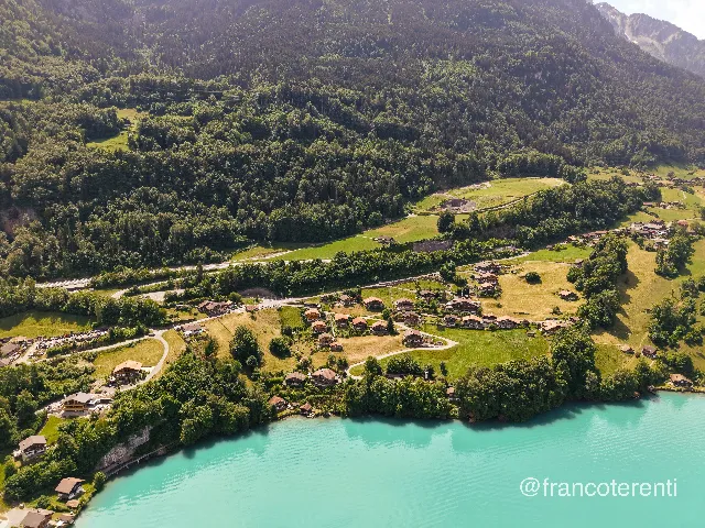 Timber chalets and mown meadows stagger down a forested mountainside toward a bright turquoise shoreline, threaded by a road, a rock cutting and scattered farm tracks under strong midday sun that leaves the upper slopes in shadow. Aerial drone photograph over ISELTWALD, Switzerland from 46.70849950999079, 7.963221073150636 on 2026-05-31. Exposure 100 · f1.8 · 9mm.