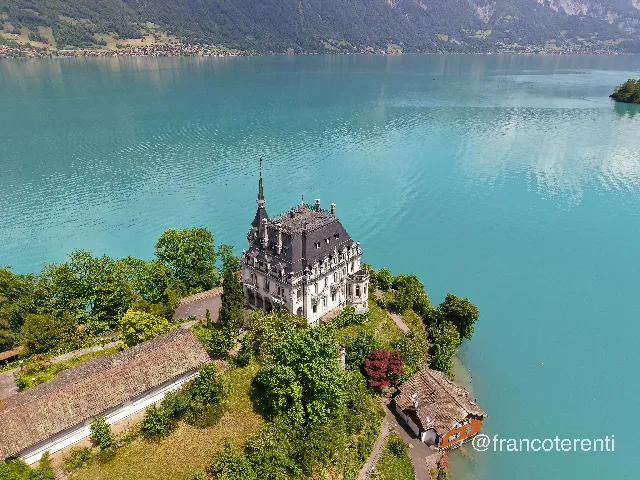 A white nineteenth-century château with a steep slate roof and slender spire stands on a wooded promontory above glassy turquoise water, flanked by a low outbuilding, a copper-leaved tree and a small orange-shuttered chalet at the water's edge, with forested slopes and a distant shore beyond. Aerial drone photograph over ISELTWALD, Switzerland from 46.71372295720992, 7.963950634002686 on 2026-05-31. Exposure 100 · f1.8 · 9mm.