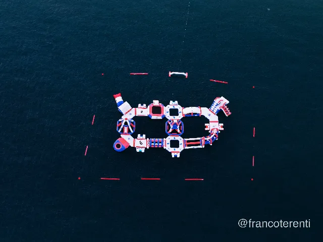 Top-down over deep blue-green open water where a red, white and blue inflatable aqua park floats slides, climbing frames, trampolines and linked platforms ringed by a wide rectangle of red marker buoys, with a single anchor line trailing off into the calm, lightly rippled surface. Aerial drone photograph over CANET DE MAR, Spain from 41.58373521535299, 2.582087538512328 on 2025-06-24. Exposure 500 · f2.8 · 19mm.