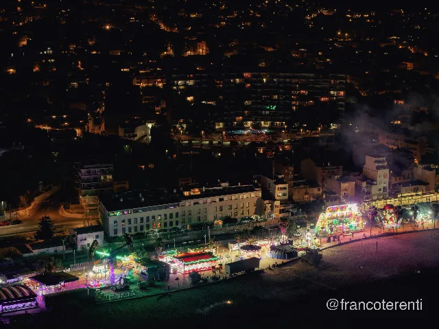 Night over a seafront fairground glowing green, red and pink — carousel, rides, food stalls and a small striped tent strung along the sand beside a lit promenade — with apartment blocks and scattered hillside windows fading into darkness and pale smoke drifting through the lights. Aerial drone photograph over CANET DE MAR, Spain from 41.58418461518206, 2.578825950622559 on 2025-06-23. Exposure 550 · f2.8 · 19mm.