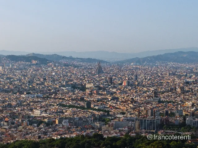Hazy golden-hour panorama across an immense low-rise city of ochre rooftops reaching back to layered blue mountain ridges, a tall spired basilica flanked by construction cranes rising above the grid, wooded slopes in the foreground and a pale cloudless sky. Aerial drone photograph over CANET DE MAR, Spain from 41.58822907289363, 2.5677537918090825 on 2025-06-25. Exposure 100 · f2.8 · 19mm.