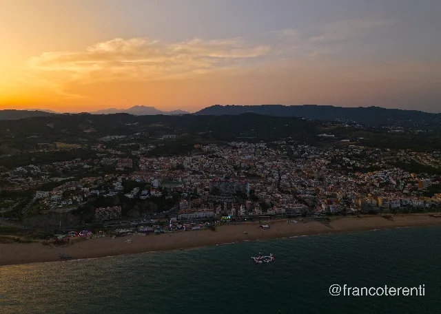 Sunset over a coastal town, orange and grey cloud banks above dark wooded hills, tightly packed pastel and terracotta buildings sloping down to a long curve of sand, bright fairground lights at the water's edge and a small inflatable park floating on the teal sea. Aerial drone photograph over CANET DE MAR, Spain from 41.586720455595746, 2.5870227813720708 on 2025-06-24. Exposure 190 · f1.8 · 9mm.