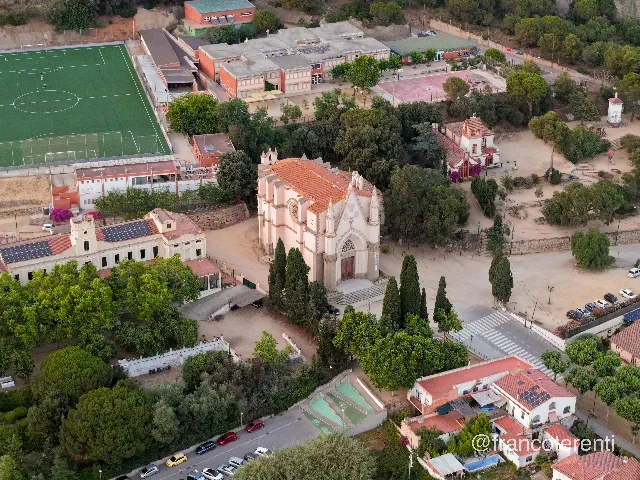 Low-angle morning light on a pink stone church with a red tiled roof and pinnacled towers, framed by tall cypresses and dusty open ground; around it a green artificial football pitch, brick school blocks, a pink hard court, solar-panelled roofs and dense stands of pine. Aerial drone photograph over CANET DE MAR, Spain from 41.59510567427761, 2.580049155624766 on 2025-06-24. Exposure 1070 · f2.8 · 19mm.