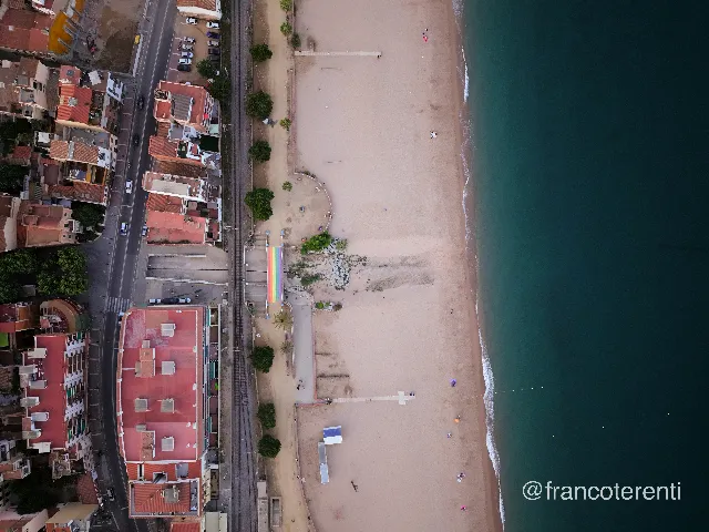 Looking straight down at the seam between a dense old town of terracotta rooftops and a broad pale-sand beach, with a railway line, palm-lined promenade and a rainbow-painted ramp running between them, and turquoise water breaking in a thin white line along the shore. Aerial drone photograph over CANET DE MAR, Spain from 41.586561111111116, 2.5825194444444444 on 2025-06-24. Exposure 640 · f1.8 · 9mm.