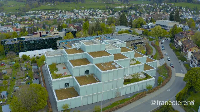 The pale glass SQUARE learning centre sits mid-frame, its stacked translucent boxes wrapped around planted green roofs and small sunken courtyards, with the dark-framed university library block and the low sports hall behind it. Allotment plots and mature trees fill the left edge, a curving road with parked cars the right, and a dense hillside town of pitched roofs and church spires recedes into flat overcast light. Aerial drone photograph over ST GALLEN, Switzerland from 47.42884211107548, 9.375522136688234. Exposure 100 · f2.8.