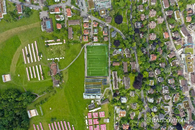 Straight down over a green hillside meadow where rows of red and white striped festival tents line up beside a farmstead with a rust coloured roof. Below them a full size artificial football pitch and a long hall roofed in solar panels give way to a leafy quarter of villas, winding streets and dark copper beeches. Aerial drone photograph over ST GALLEN, Switzerland from 47.427492084392746, 9.372003078460695 on 2024-05-30. Exposure 100 · f4 · 10mm.